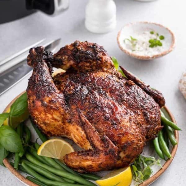 Whole air fryer rotisserie chicken set on a platter with herbs, green beans, and lemon wedges. An air fryer is visible in the background.