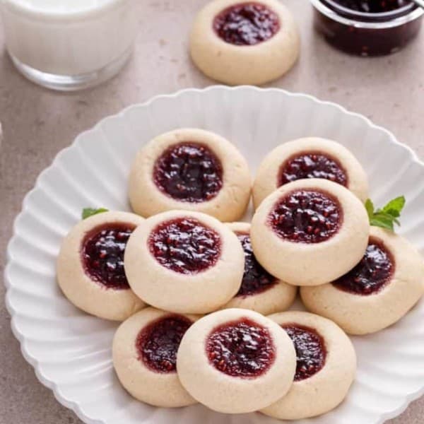 Raspberry thumbprint cookies piled onto a white plate. A glass of milk and a bowl of raspberry jam are visible in the background.