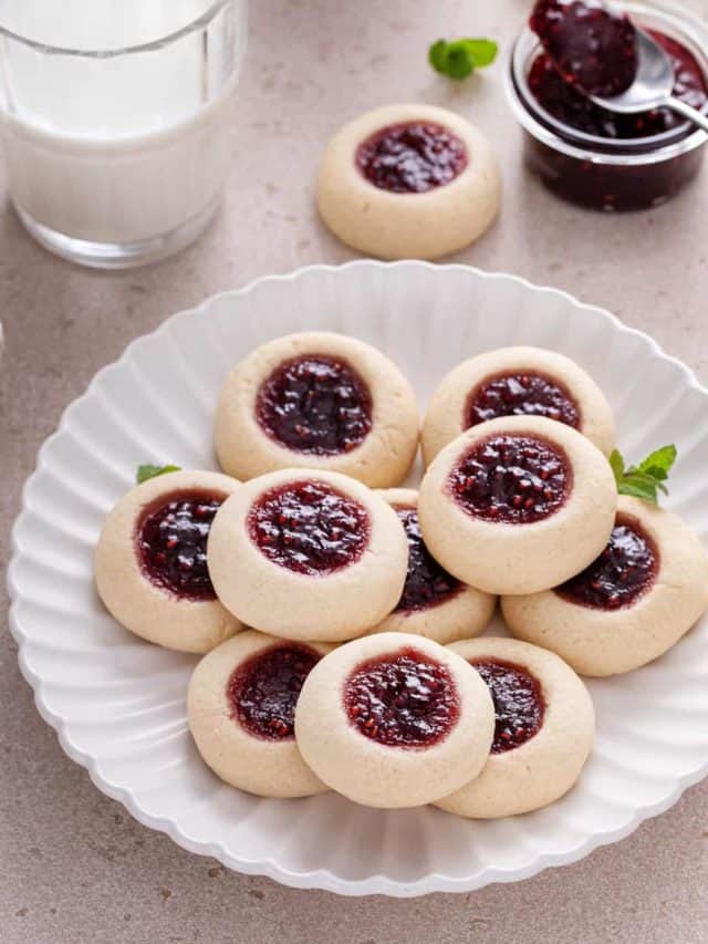 Raspberry thumbprint cookies piled onto a white plate. A glass of milk and a bowl of raspberry jam are visible in the background.