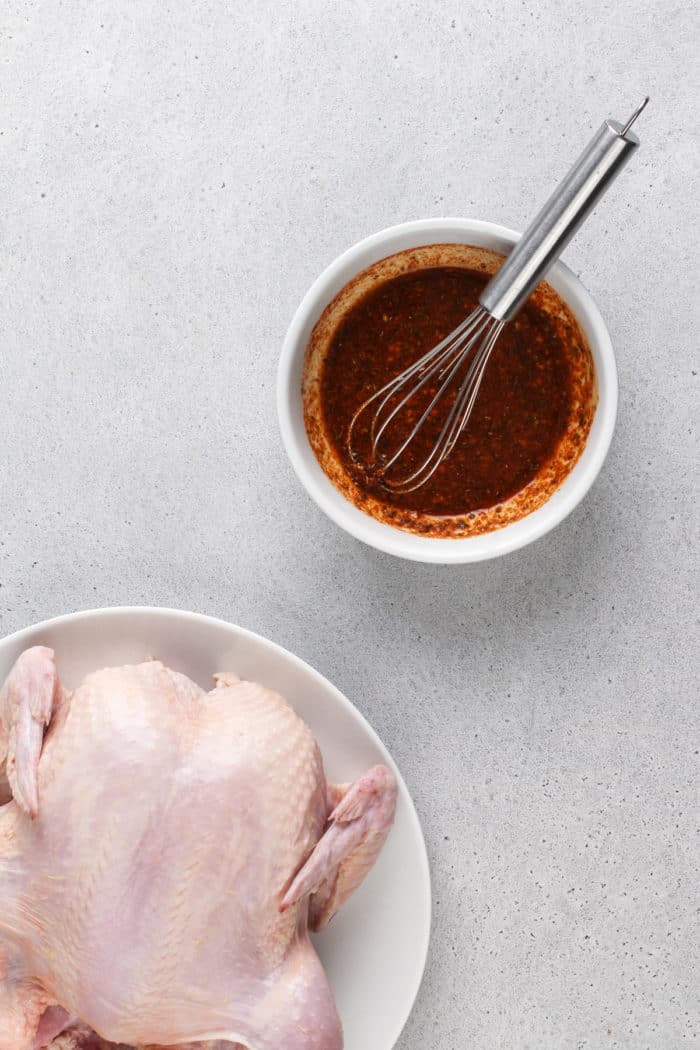 Bowl of spices mixed with olive oil next to a whole chicken on a white plate.