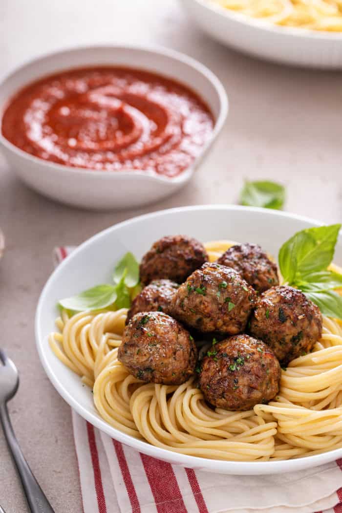 Bowl of spaghetti topped with air fryer meatballs in the foreground. A bowl of pasta sauce is in the background.
