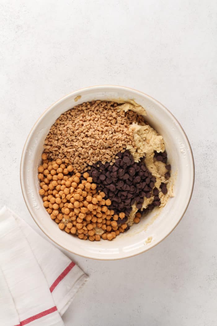 Caramel bits, toffee bits, and chocolate chips being added to cookie dough in a ceramic bowl.