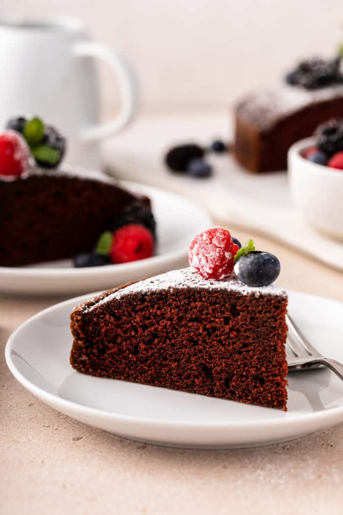 Slice of eggless chocolate cake, topped with powdered sugar and berries, on a white plate.