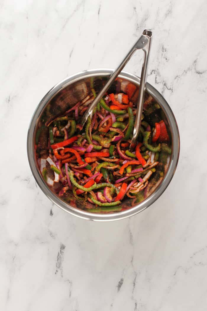 Tongs tossing sliced peppers and onions together with fajita spices in a metal mixing bowl.