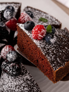 Cake server holding up a slice of eggless chocolate cake, dusted in powdered sugar and topped with fresh berries.