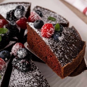 Cake server holding up a slice of eggless chocolate cake, dusted in powdered sugar and topped with fresh berries.