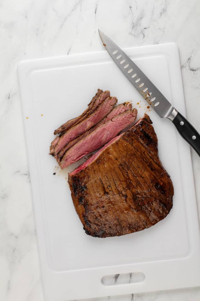 Grilled and marinated flank steak being sliced on a white cutting board.