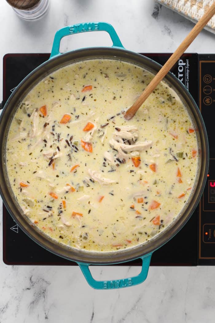 Wooden spoon stirring a large pot of chicken and wild rice soup on a burner.