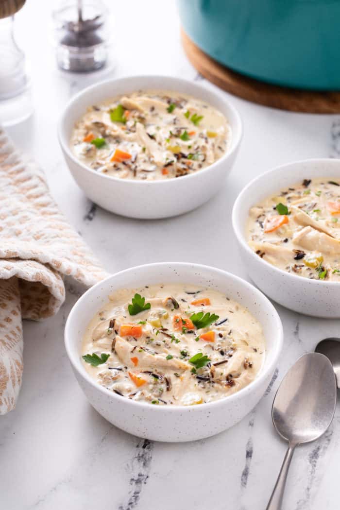 Three bowls of chicken and wild rice soup set next to a blue dutch oven on a marble countertop.