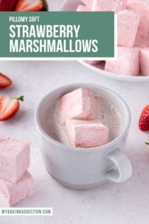 Two strawberry marshmallows in a white mug of hot chocolate. A bowl of strawberry marshmallows is in the background. Text overlay includes recipe name.