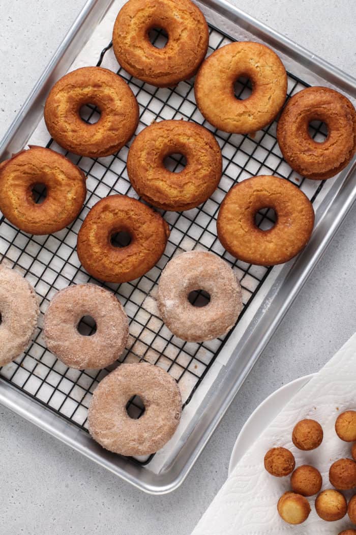 Cake donuts cooling on a wire rack set over a sheet tray. Some of the donuts are covered in cinnamon sugar.