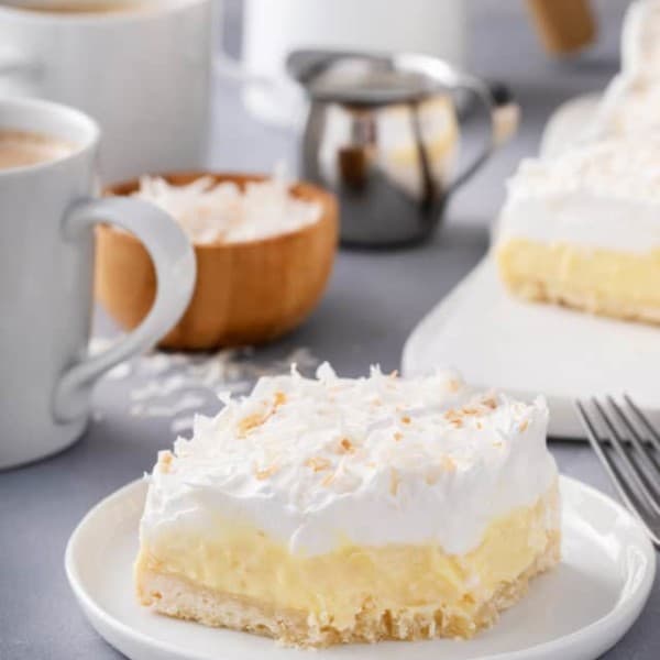 Plated coconut cream bar with cups of coffee in the background. A bite has been taken from the corner of the bar.