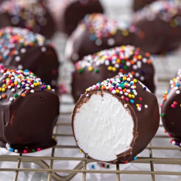 Several chocolate-covered marshmallows topped with rainbow sprinkles on a wire rack. A marshmallow in front has been cut in half to show the center.