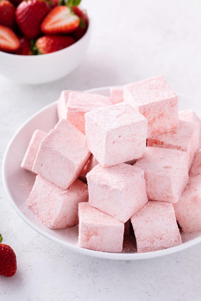 White bowl filled with strawberry marshmallows. A bowl of sliced strawberries is in the background.