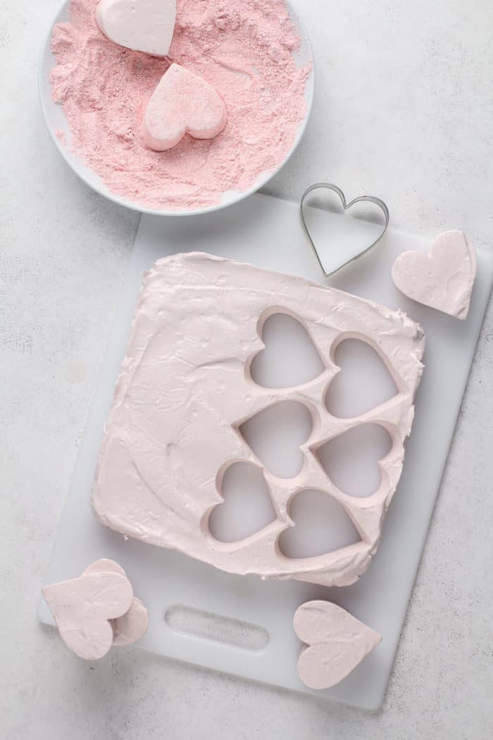 Cutting heart shapes out of a slab of strawberry marshmallows on a white cutting board.