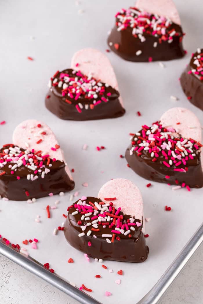 Heart-shaped strawberry marshmallows halfway dipped in chocolate and topped with festive sprinkles set on a parchment-lined baking sheet.