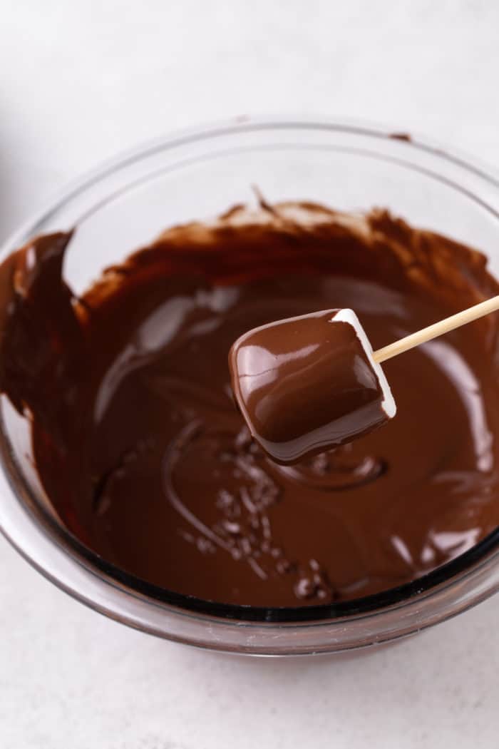 Marshmallow on a wooden skewer being dipped into a bowl of melted chocolate.