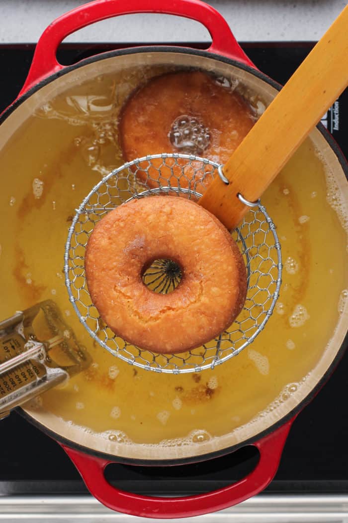Fried cake donut being lifted out of the frying oil with a wire spider.