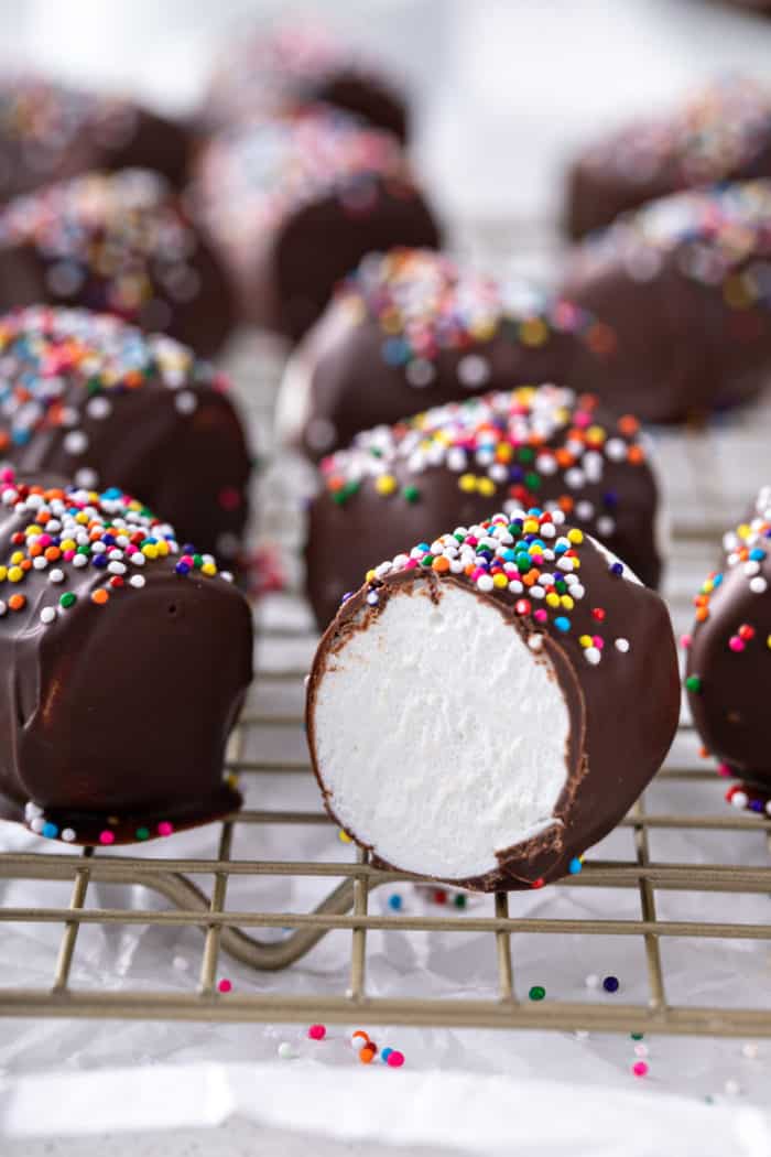 Several chocolate-covered marshmallows topped with rainbow sprinkles on a wire rack. A marshmallow in front has been cut in half to show the center.
