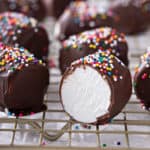 Several chocolate-covered marshmallows topped with rainbow sprinkles on a wire rack. A marshmallow in front has been cut in half to show the center.