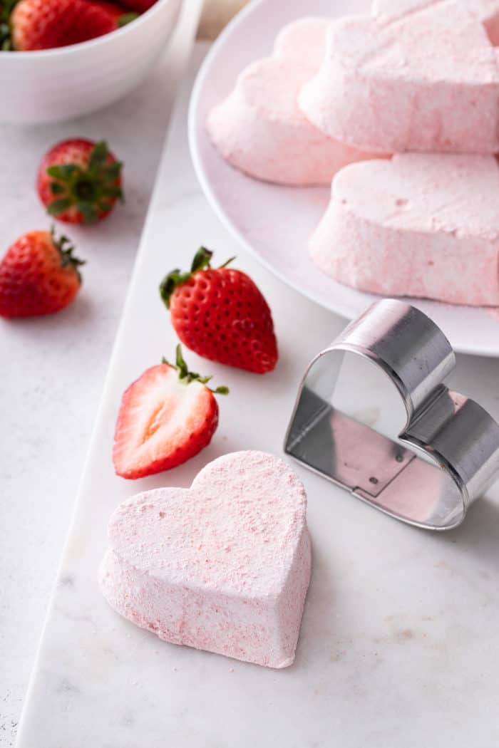 Heart-shaped strawberry marshmallow next to a metal heart cookie cutter on a white board.