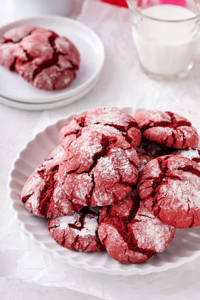 White plate filled with red velvet crinkle cookies. A glass of milk is visible in the background.