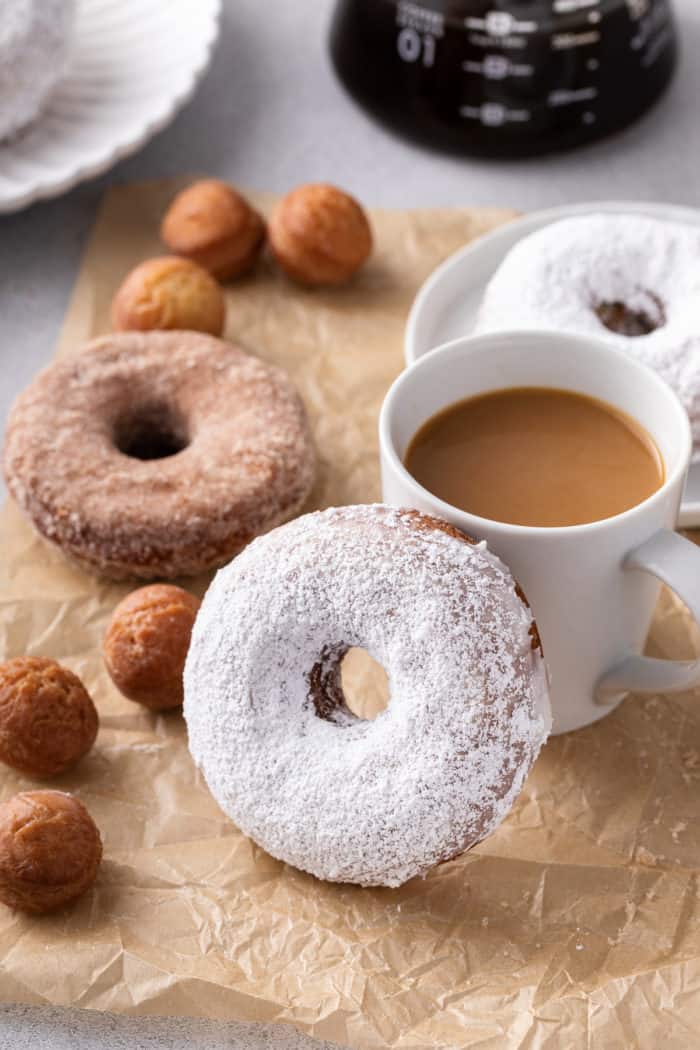 Powdered-sugar-coated cake donut propped up next to a cup of coffee. More cake donuts and donut holes are visible in the background.