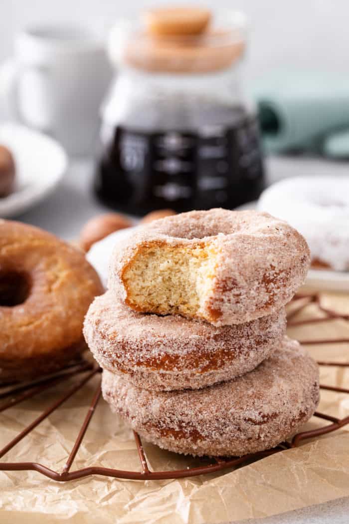 Three cake donuts covered in cinnamon sugar stacked on a wire rack. The top donut has a bite taken from it.