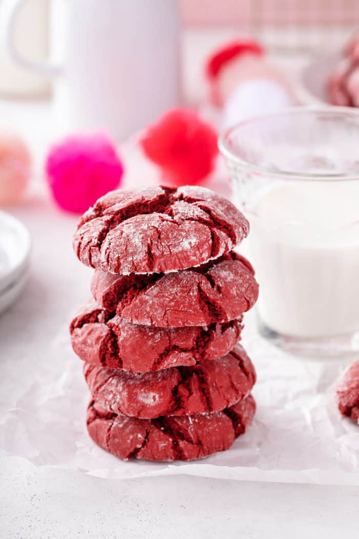 Five red velvet crinkle cookies stacked on a piece of parchment in front of a glass of milk.