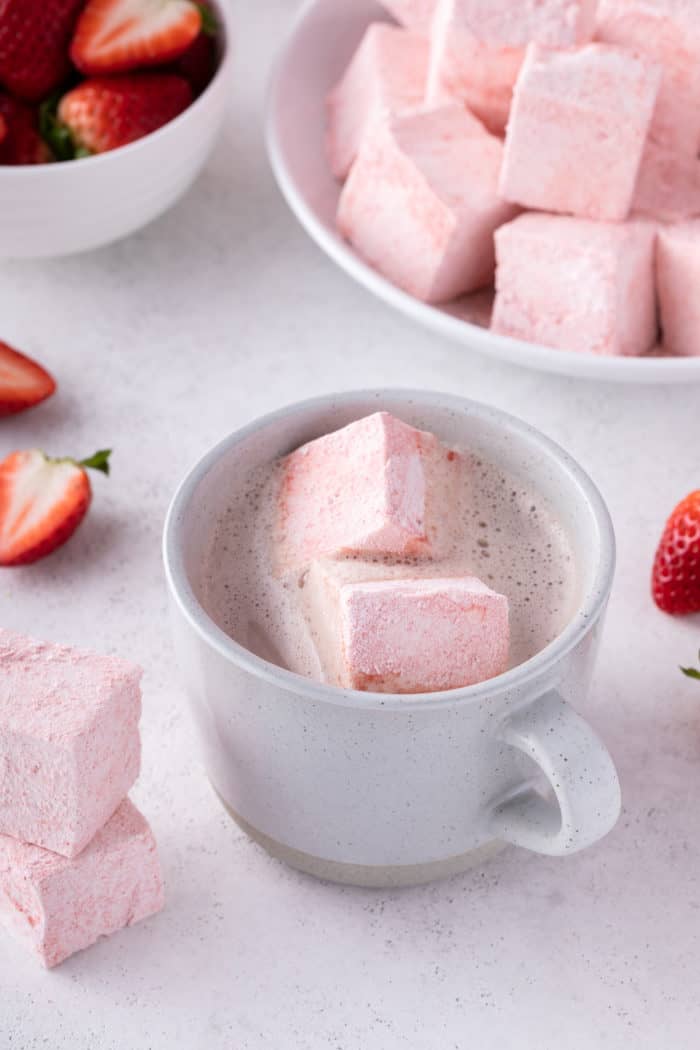 Two strawberry marshmallows in a white mug of hot chocolate. A bowl of strawberry marshmallows is in the background.