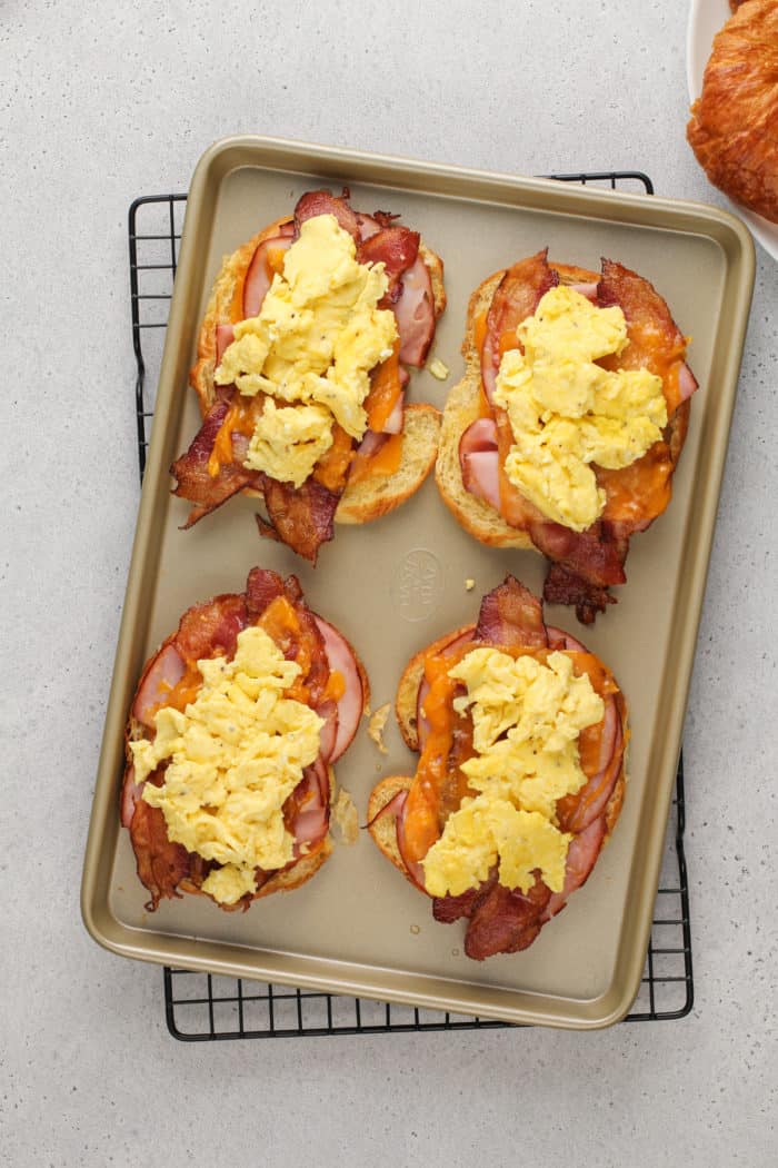 Eggs added onto warmed croissant breakfast sandwiches on a sheet pan.