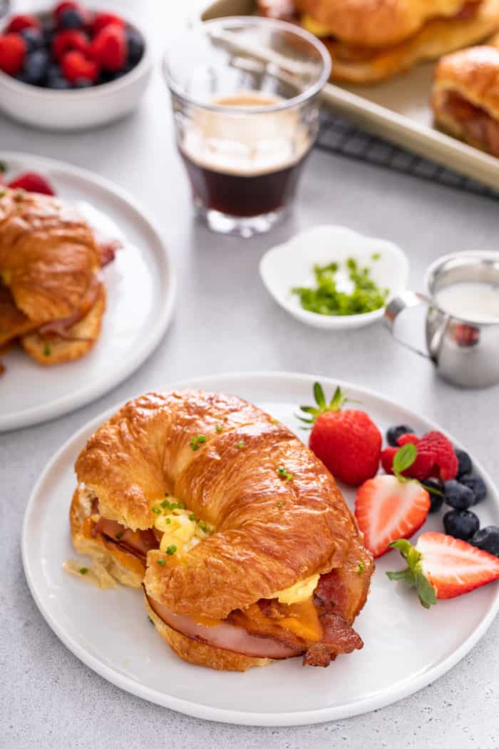 Croissant breakfast sandwich next to fresh berries on a white plate. A second plate and more fruit is visible in the background.