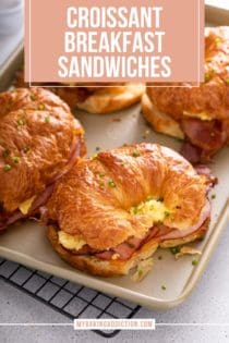 Assembled and baked croissant breakfast sandwiches on a sheet pan, topped with fresh chives. Text overlay includes recipe name.