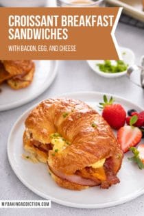 Croissant breakfast sandwich next to fresh berries on a white plate. A second plate and more fruit is visible in the background. Text overlay includes recipe name.