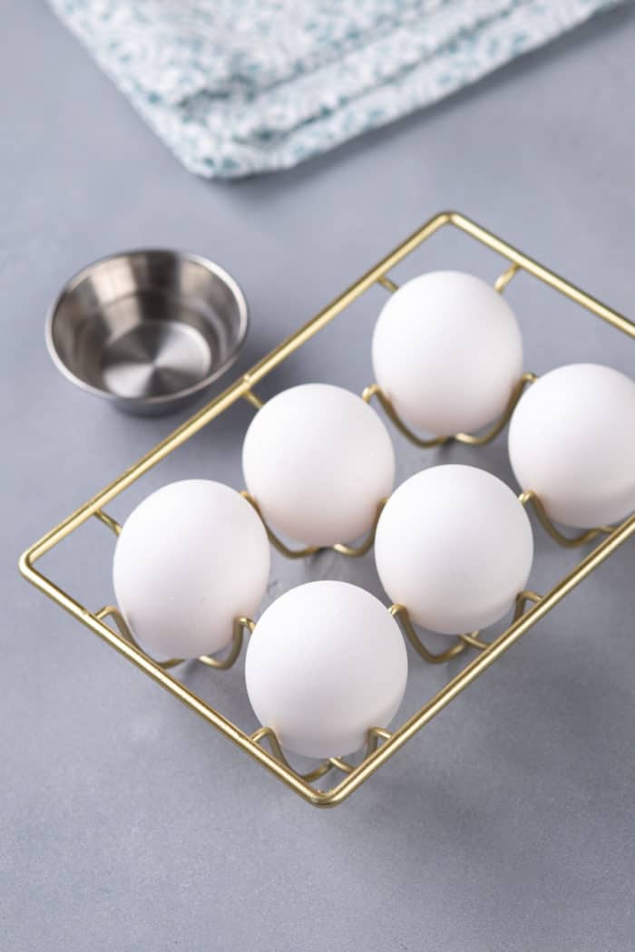 Six whole eggs and a small bowl of white vinegar on a countertop.