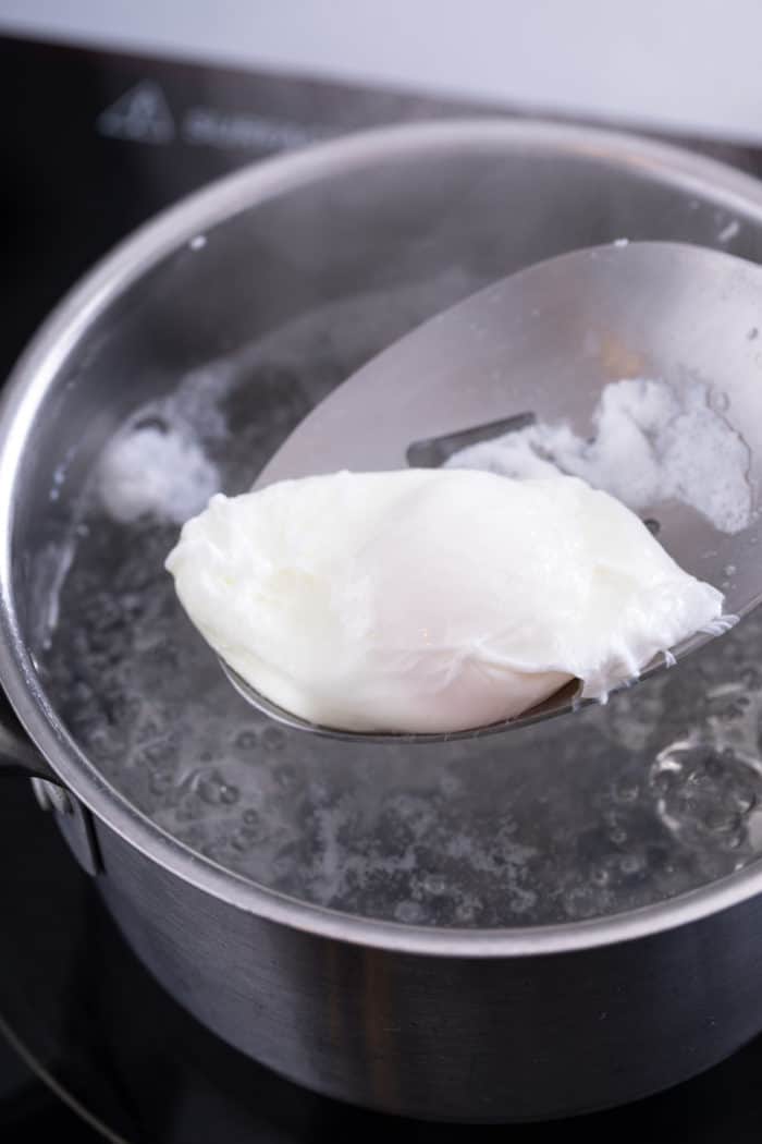 Freshly poached egg being removed from simmering water with a slotted spoon.