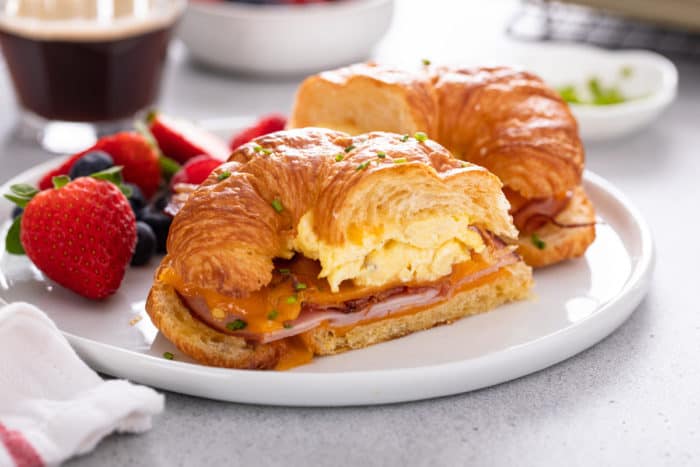 Halved croissant breakfast sandwich on a white plate next to fresh berries.