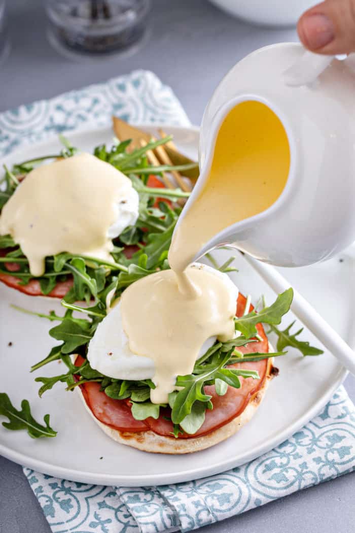 Hollandaise sauce being poured over eggs benedict on a white plate.