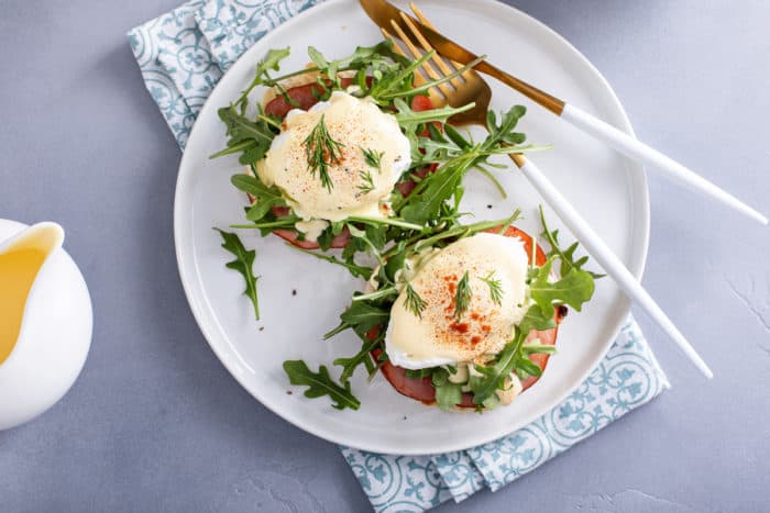 Overhead view of eggs benedict next to a fork and knife on a white plate, set on a blue napkin.