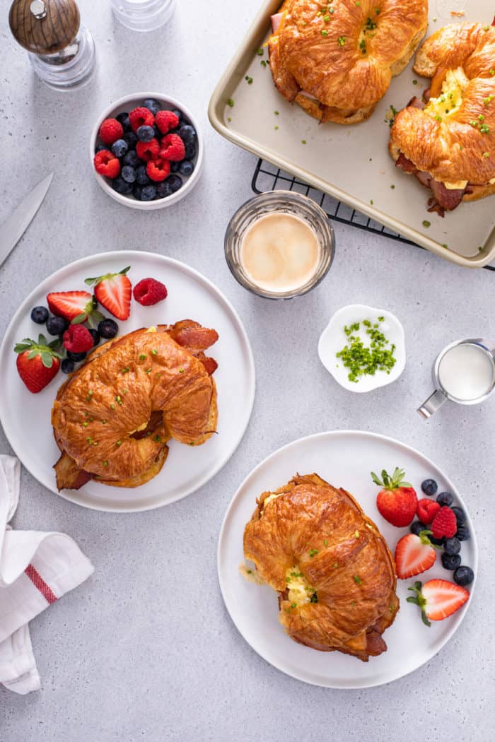 Overhead view of two white plates, each filled with a croissant breakfast sandwich and fresh fruit.