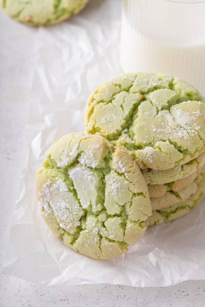 Several stacked green crinkle cookies next to a glass of milk. One of the cookies is leaning against the stack.