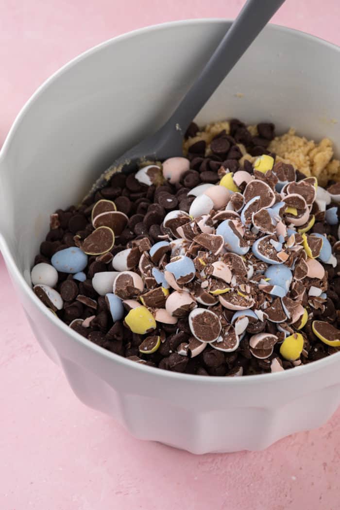 Chocolate chips and chopped cadbury mini eggs being added to a white mixing bowl of cookie dough.