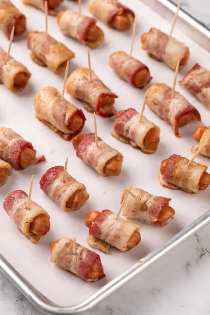 Little smokies wrapped in bacon and set on a parchment-lined baking sheet, ready to go in the oven.
