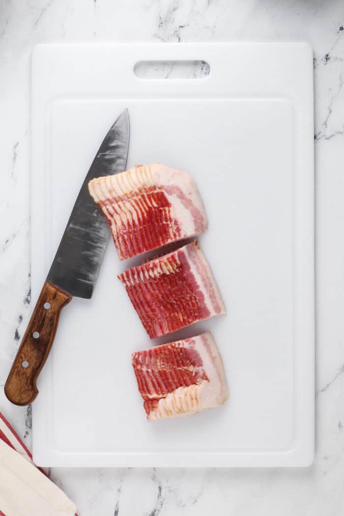 Slab of bacon cut into 3 equal sections on a white cutting board, next to a chef's knife.