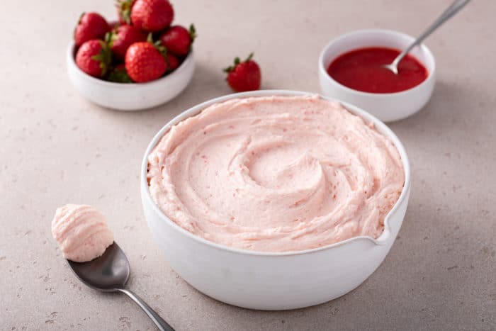 White bowl filled with strawberry frosting set on a countertop next to fresh strawberries and a small bowl of strawberry puree.