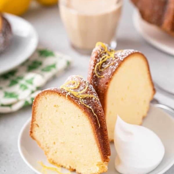 Two slices of lemon cream cheese pound cake garnished with lemon zest and a dollop of whipped cream on a white plate.