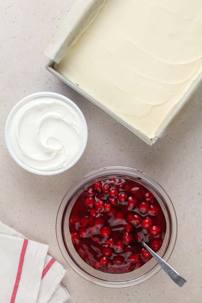Pan of nana's easy cheesecake next to bowls of whipped topping and cherry pie filling.
