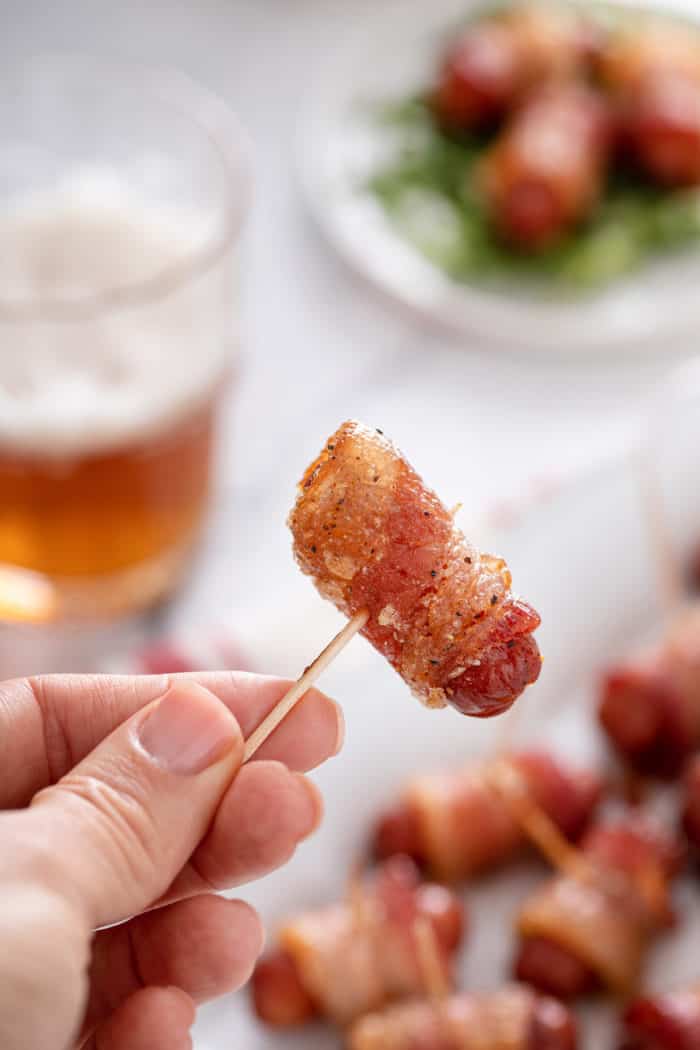 Hand holding up a bacon-wrapped little smokie on a toothpick.