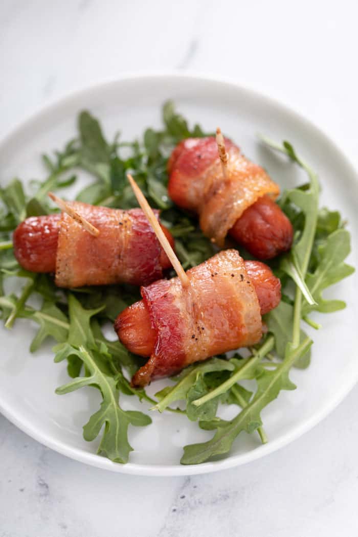 Three bacon-wrapped little smokies on a bed of arugula on a small white plate.