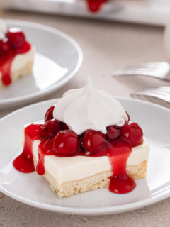 Slice of nana's easy cheesecake, topped with cherry pie filling and whipped cream, on a white plate.
